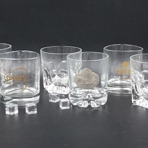 Peut inclure: Ensemble de six verres à whisky transparents. Certains verres présentent le logo "Grant's" en lettres dorées, tandis que d'autres arborent un emblème "Whisky" argenté. Des glaçons transparents sont disposés devant les verres.