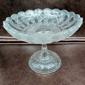 May include: A clear glass pedestal bowl with a decorative, scalloped edge and a raised, patterned base. The bowl is perfect for serving desserts, snacks, or fruit.