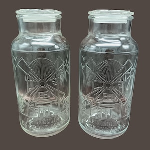 Può includere: Due barattoli di vetro trasparente con coperchi, ognuno con un disegno inciso di un mulino a vento con il testo "Le Moulin de Grand-Mere" in basso.