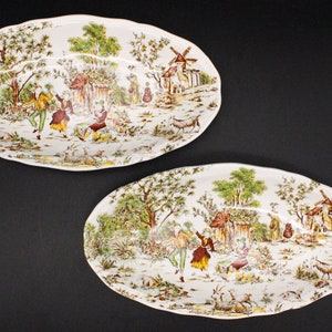 Puede incluir: Dos platos de servir ovalados blancos con un diseño de escena campestre marrón y verde. La escena presenta un molino de viento, una cabaña y personas bailando en un campo.