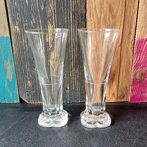 Peut inclure: Deux verres à boire clairs à pied. Les verres ont un dessus évasé et une base décorative. Les verres sont en verre transparent et sont vides. L'arrière-plan est un mélange de planches de bois de différentes couleurs.