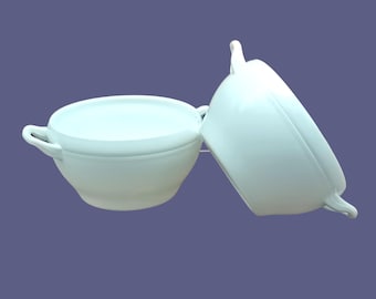 May include: Two white ceramic soup bowls with handles and lids. One bowl is upright, and the other is tilted on its side. The bowls have a smooth, glossy finish and are set against a muted purple background.