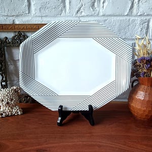 May include: An octagonal serving platter with a white center and a border featuring black parallel lines arranged in a geometric pattern. The platter is supported by a black plastic stand, showcasing a clean design with sharp, contrasting stripes.