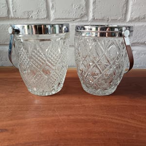 Puede incluir: Dos cubos de hielo de vidrio transparente con bordes y asas plateadas. Los cubos tienen un patrón de diamantes y asas de cuero marrón. Los cubos están sobre una superficie de madera.