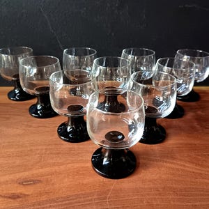 May include: A set of ten clear glass cordial glasses with black stems and bases. The glasses are arranged on a wooden surface, with a dark background. The glasses have a rounded bowl shape.