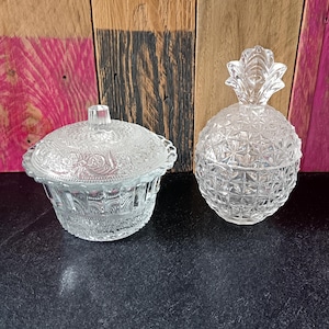 Könnte beinhalten: Zwei Bonbonnieren aus klarem Glas. Eine ist rund mit Deckel und dekorativem Rand. Die andere ist wie eine Ananas mit Deckel geformt. Beide Schalen stehen auf einer dunklen Oberfläche mit Holzgrund.