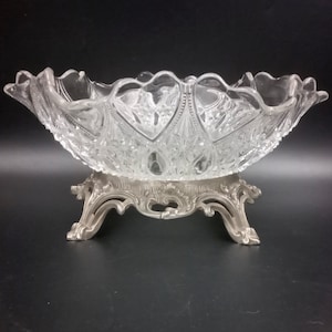 May include: A clear glass bowl with a cut glass design sits on a silver stand with three legs. The bowl is oval shaped and has a scalloped edge.