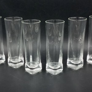 Peut inclure: Six verres à boire en verre transparent avec une base carrée. Les verres sont vides et disposés en rangée sur une surface noire.