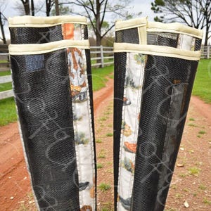 May include: Pair of black mesh boot covers with a cream-colored trim. The center panel features a patterned fabric with cowboy boots, hats, and other western-themed imagery. The covers are designed to protect boots and are set on a dirt path.