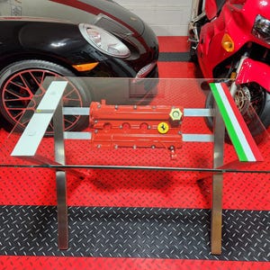 May include: A glass-top coffee table with a red Ferrari engine block as its centerpiece. The table has a stainless steel frame and features white and Italian flag accents. The table is displayed on a red and black diamond-plate floor.