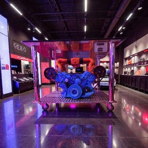 May include: A blue engine with orange piping is displayed on a chrome cart with wheels. The cart is in a showroom with dark walls and bright overhead lights. The floor is a dark, reflective material.
