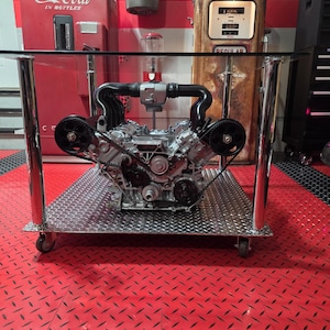 Könnte beinhalten: Ein einzigartiger Couchtisch mit einem silbernen Motorblock als Basis, gekrönt von einer transparenten Glasoberfläche. Der Tisch hat ein Riffelblechregal und verchromte Beine mit Rollen. Im Hintergrund befinden sich ein Coca-Cola-Automat und eine Zapfsäule.