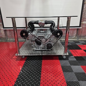 Könnte beinhalten: Ein einzigartiger Couchtisch mit einem Automotor als Basis. Der silberne Motor mit schwarzen Komponenten steht auf einem Riffelblech-Metallregal. Der Tisch hat eine transparente Glasplatte und Chrombeine. Der Boden ist rot und schwarz.