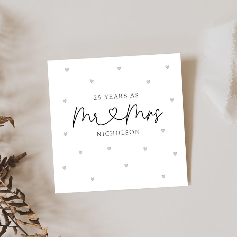 Op de afbeelding: Een witte kaart met zwarte tekst die "25 Years as Mr & Mrs Nicholson" luidt, met een klein hartje tussen de woorden "Mr" en "Mrs". De kaart is versierd met kleine grijze hartjes.