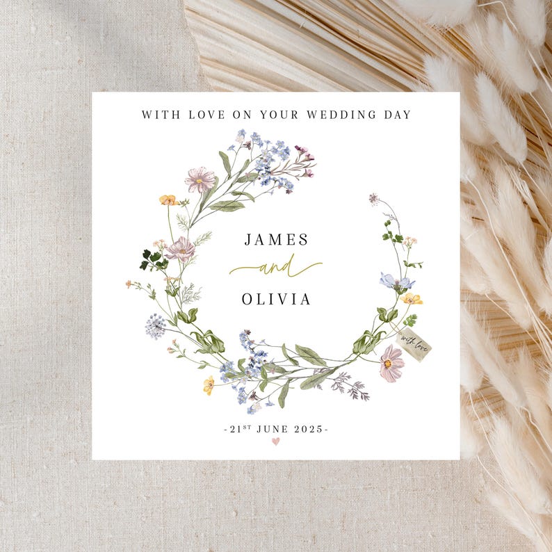 May include: A white card with a floral wreath design. The wreath is made of watercolor illustrations of various flowers and greenery. The text "With love on your wedding day" is at the top of the card. The names "James and Olivia" are in the center of the wreath, with the date "21st June 2025" below.