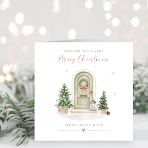 Op de afbeelding: Een vierkante kerstkaart met een aquarelillustratie van een boogvormige saliegroene deur met een krans, geflankeerd door twee kleine dennenbomen en een lantaarn. De tekst luidt: WISHING YOU A VERY Merry Christmas, JAMES, OLIVIA & FIN.