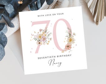 May include: A white greeting card with a pink "70" and floral accents. The text reads "With Love On Your", "Seventieth Birthday", and "Nanny". The card is decorated with delicate flowers and leaves.