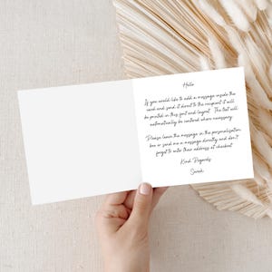 Puede incluir: Una tarjeta de felicitaci&oacute;n blanca abierta, con texto manuscrito en tinta negra. El texto proporciona instrucciones para agregar un mensaje y enviarlo directamente al destinatario. La tarjeta est&aacute; sostenida por una mano, sobre un fondo neutro.
