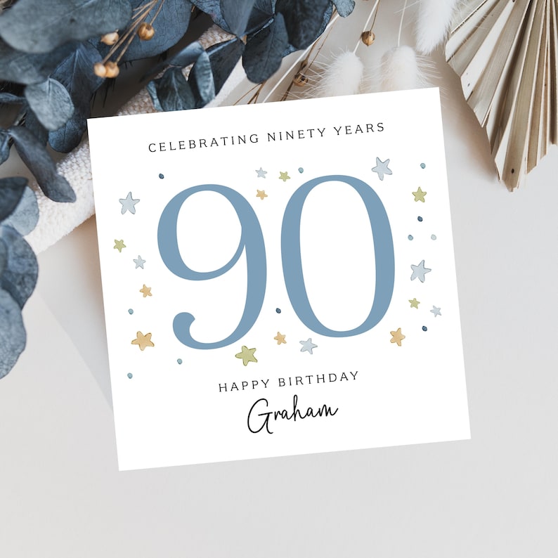 Puede incluir: Tarjeta de cumplea&ntilde;os blanca con el n&uacute;mero 90 en azul, rodeada de ilustraciones de estrellas. La tarjeta dice "Celebrating Ninety Years" y "Happy Birthday Graham" en escritura negra. Elementos decorativos visibles en el fondo.
