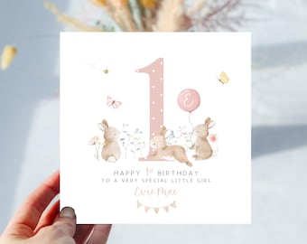 May include: A birthday card with a pink number 1, bunnies, and butterflies. The card reads "Happy 1st Birthday to a very special little girl" with a name below. A pink balloon with a letter floats above the bunnies.