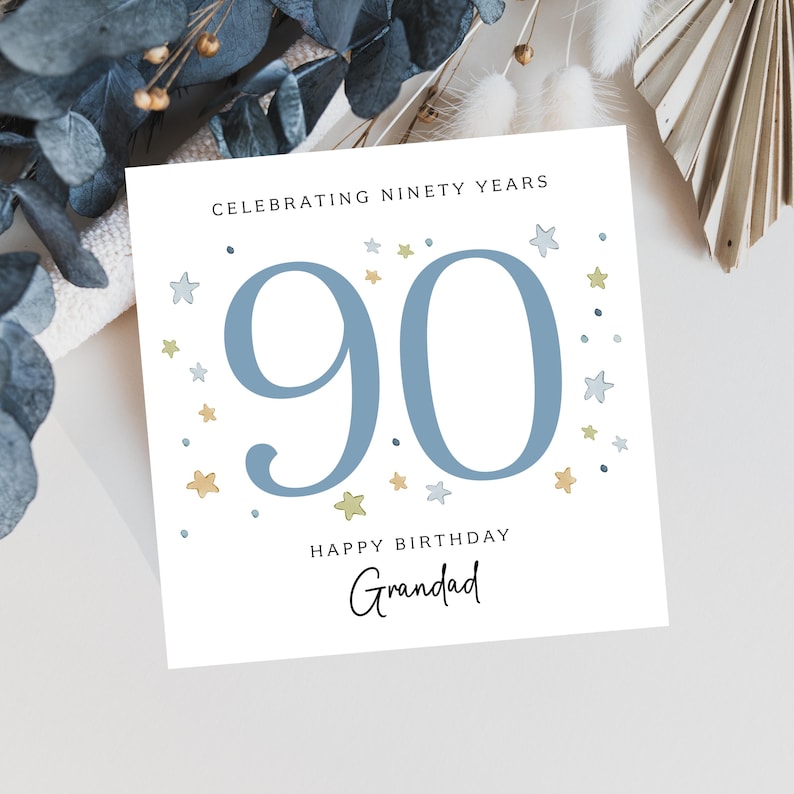 Puede incluir: Tarjeta de cumplea&ntilde;os blanca con el n&uacute;mero 90 en azul, rodeada de peque&ntilde;as estrellas. La tarjeta dice "Celebrating Ninety Years" y "Happy Birthday Grandad" en escritura elegante.