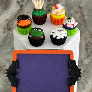 Op de afbeelding: Zes Halloween-thema cupcakes met verschillende decoraties, waaronder een spin, een hand, een oog, een mummie, ma&iuml;s en een monster. De cupcakes zijn gerangschikt op een wit oppervlak met een paarse krijtbord met een zwarte vleermuis aan elke kant.