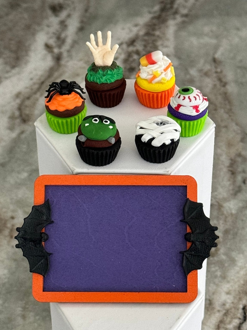 Op de afbeelding: Zes Halloween-thema cupcakes met verschillende decoraties, waaronder een spin, een hand, een oog, een mummie, ma&iuml;s en een monster. De cupcakes zijn gerangschikt op een wit oppervlak met een paarse krijtbord met een zwarte vleermuis aan elke kant.