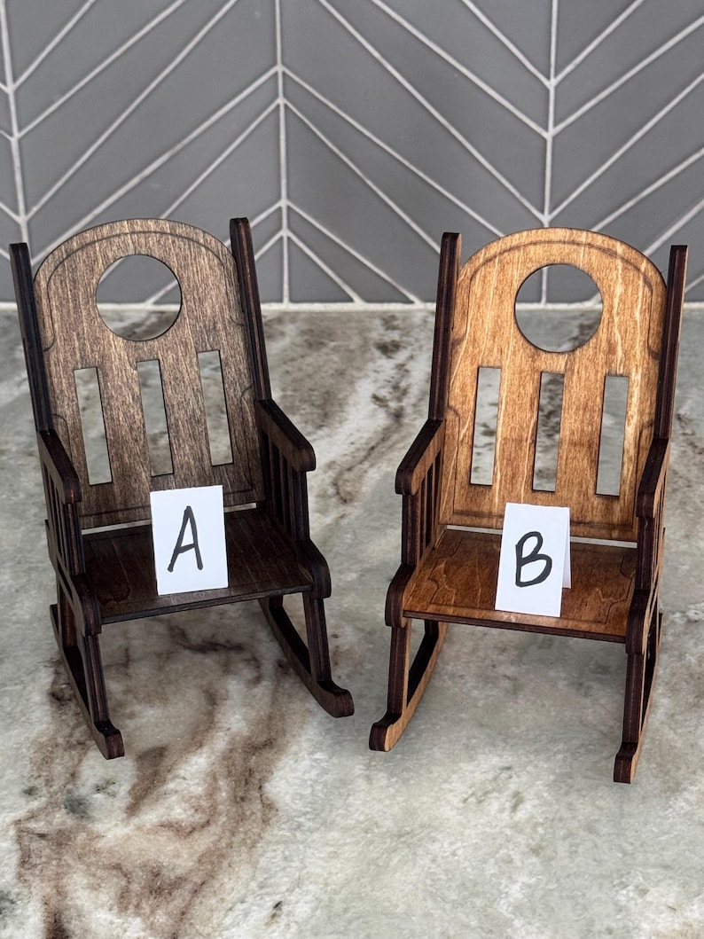 Peut inclure: Deux petites chaises &agrave; bascule en bois, une marron fonc&eacute; et une marron clair, sont pr&eacute;sent&eacute;es c&ocirc;te &agrave; c&ocirc;te. Chaque chaise a un dossier arrondi avec une d&eacute;coupe circulaire et des lattes verticales. De petites cartes blanches &eacute;tiquet&eacute;es "A" et "B" sont plac&eacute;es sur les si&egrave;ges.