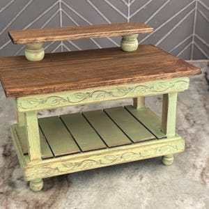 May include: A small, two-tiered wooden table with a distressed finish. The top tier is a dark brown, while the base and lower shelf are painted a light green. Decorative leaf carvings adorn the edges. The table is approximately 12 inches long.