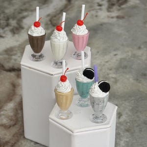 May include: Miniature milkshakes in chocolate, vanilla, pink, teal, gold, and silver. Each milkshake has whipped cream, a cherry, and a straw. Some milkshakes have an Oreo cookie. The milkshakes are displayed on white hexagonal pedestals.
