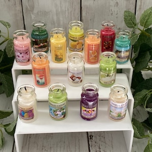 May include: A collection of 14 small glass candle jars with various scents and labels. The candles are arranged on a white tiered shelf. The candles are all in different colors, including red, green, blue, yellow, and orange. The labels on the candles include names like "Christmas Garland", "Ocean Lemon", "Vanilla Lime", and "Happy Easter".
