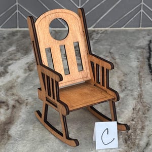 Peut inclure: Une chaise ber&ccedil;ante miniature en bois avec une finition brun clair. La chaise a un dossier arrondi avec une d&eacute;coupe circulaire et des lattes verticales. L'assise et les accoudoirs sont &eacute;galement visibles. Une petite carte blanche avec la lettre "C" est devant la chaise.