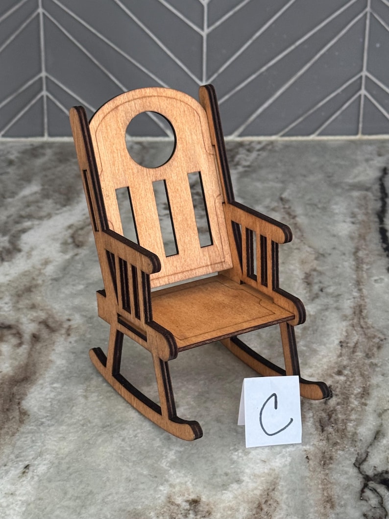 Peut inclure: Une chaise ber&ccedil;ante miniature en bois avec une finition brun clair. La chaise a un dossier arrondi avec une d&eacute;coupe circulaire et des lattes verticales. L'assise et les accoudoirs sont &eacute;galement visibles. Une petite carte blanche avec la lettre "C" est devant la chaise.
