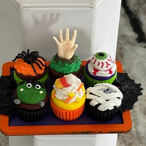 Op de afbeelding: Zes Halloween-cupcakes met verschillende griezelige decoraties, waaronder een spin, een hand, een oog, candy corn en een mummie. De cupcakes zijn gerangschikt op een zwart en oranje dienblad.