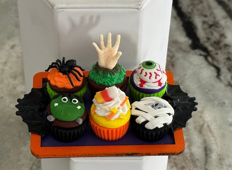 Op de afbeelding: Zes Halloween-cupcakes met verschillende griezelige decoraties, waaronder een spin, een hand, een oog, candy corn en een mummie. De cupcakes zijn gerangschikt op een zwart en oranje dienblad.
