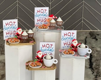 May include: Miniature holiday displays featuring a sign that reads HOT chocolate is like a hug from the inside. Each set includes a small Santa Claus or gingerbread figure, a mug of hot cocoa with whipped cream, and a plate of cookies.