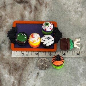Op de afbeelding: Een set van zes Halloween-cupcakes versierd met verschillende griezelige ontwerpen, waaronder een oogbal, een mummie, een monster, een spin en een ma&iuml;s. De cupcakes zijn gerangschikt op een paarse dienblad met een zwart vleermuisontwerp.