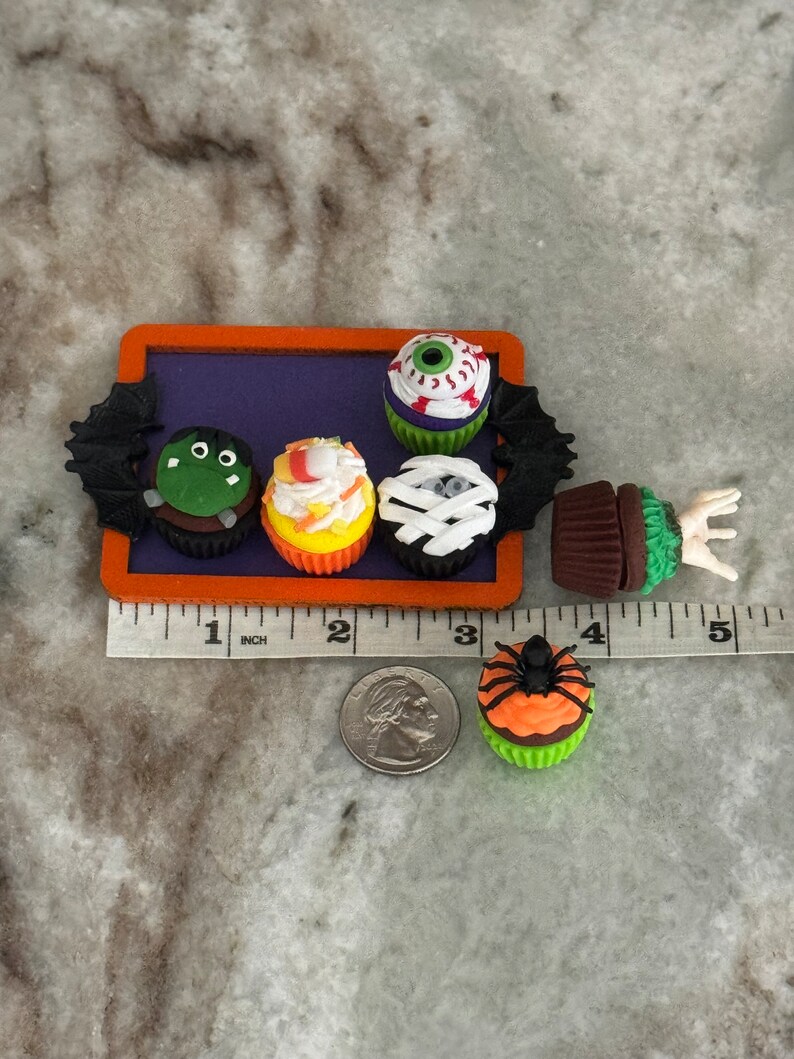 Op de afbeelding: Een set van zes Halloween-cupcakes versierd met verschillende griezelige ontwerpen, waaronder een oogbal, een mummie, een monster, een spin en een ma&iuml;s. De cupcakes zijn gerangschikt op een paarse dienblad met een zwart vleermuisontwerp.