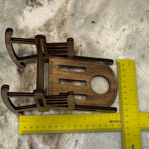 Peut inclure: Petit tra&icirc;neau jouet en bois, d'environ 13 cm de long, avec une finition marron fonc&eacute;. Le tra&icirc;neau a un design classique avec des patins incurv&eacute;s et un si&egrave;ge &agrave; lattes. Le tra&icirc;neau est pr&eacute;sent&eacute; sur une surface claire.