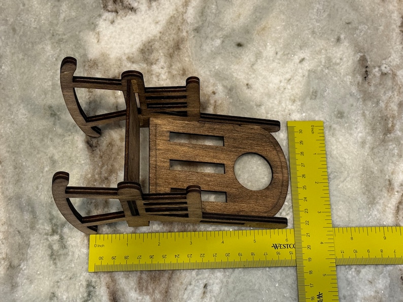 Peut inclure: Petit tra&icirc;neau jouet en bois, d'environ 13 cm de long, avec une finition marron fonc&eacute;. Le tra&icirc;neau a un design classique avec des patins incurv&eacute;s et un si&egrave;ge &agrave; lattes. Le tra&icirc;neau est pr&eacute;sent&eacute; sur une surface claire.