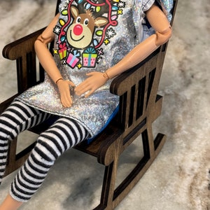 Peut inclure: Une poup&eacute;e v&ecirc;tue d'un haut argent&eacute; &agrave; paillettes avec un motif de renne et d'un legging ray&eacute; noir et blanc. La poup&eacute;e est assise dans un petit rocking-chair en bois. Le motif du renne comprend un nez rouge, des lumi&egrave;res color&eacute;es et des bo&icirc;tes cadeaux.