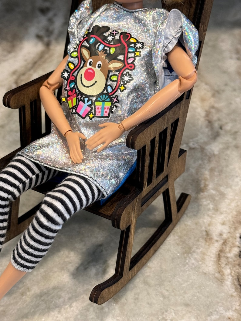 Peut inclure: Une poup&eacute;e v&ecirc;tue d'un haut argent&eacute; &agrave; paillettes avec un motif de renne et d'un legging ray&eacute; noir et blanc. La poup&eacute;e est assise dans un petit rocking-chair en bois. Le motif du renne comprend un nez rouge, des lumi&egrave;res color&eacute;es et des bo&icirc;tes cadeaux.