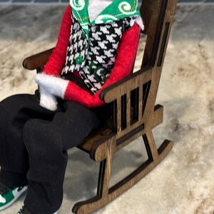 Peut inclure: Une figurine miniature v&ecirc;tue d'une chemise rouge &agrave; manches longues, d'un pantalon noir et de gants blancs, assise dans un rocking-chair en bois. La figurine porte un gilet pied-de-poule et une &eacute;charpe verte et blanche. La figurine porte des baskets noires et blanches avec des accents verts.