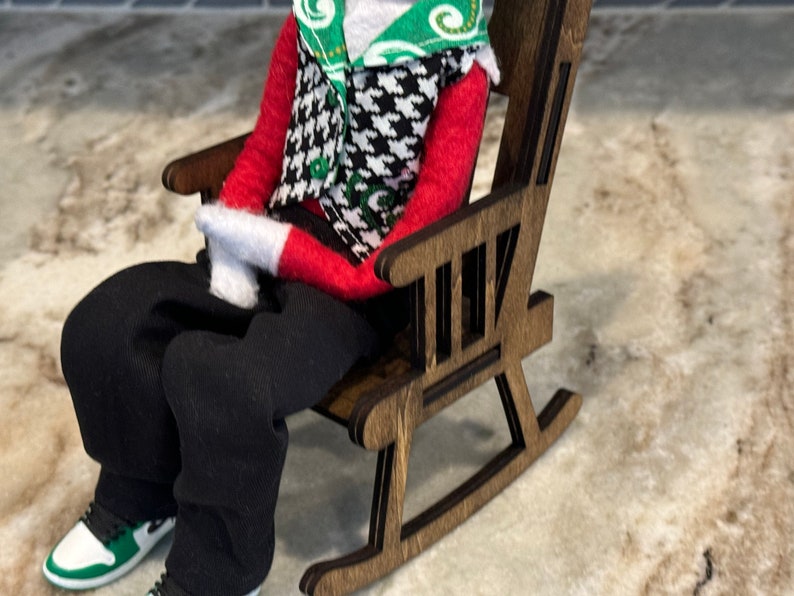 Peut inclure: Une figurine miniature v&ecirc;tue d'une chemise rouge &agrave; manches longues, d'un pantalon noir et de gants blancs, assise dans un rocking-chair en bois. La figurine porte un gilet pied-de-poule et une &eacute;charpe verte et blanche. La figurine porte des baskets noires et blanches avec des accents verts.