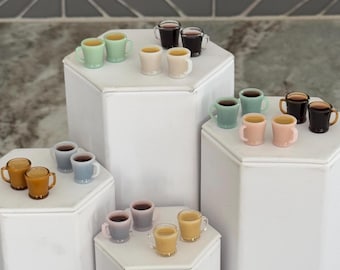 May include: A collection of miniature ceramic mugs in various colors, including light blue, light green, pink, and brown. The mugs are arranged on three different white platforms.