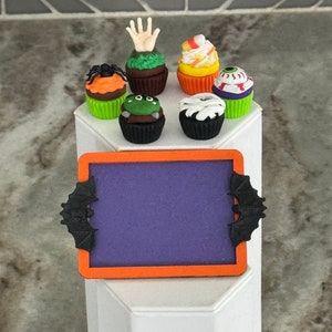 Op de afbeelding: Een paars schoolbord met een zwarte vleermuis aan elke kant, dat op een wit voetstuk staat. Zes Halloween-cupcakes met verschillende decoraties, waaronder een hand, een spin, een oog en ma&iuml;s, zijn rond het schoolbord gerangschikt.