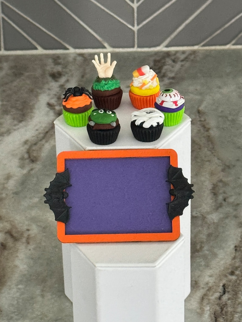 Op de afbeelding: Een paars schoolbord met een zwarte vleermuis aan elke kant, dat op een wit voetstuk staat. Zes Halloween-cupcakes met verschillende decoraties, waaronder een hand, een spin, een oog en ma&iuml;s, zijn rond het schoolbord gerangschikt.