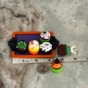 Op de afbeelding: Een set van zes Halloween-thema cupcakes met verschillende griezelige decoraties, waaronder een oog, een spin, een monster en snoep ma&iuml;s. De cupcakes zijn gerangschikt op een paarse dienblad met een zwart vleermuisontwerp. De dienblad staat op een wit aanrecht met een liniaal voor schaal.