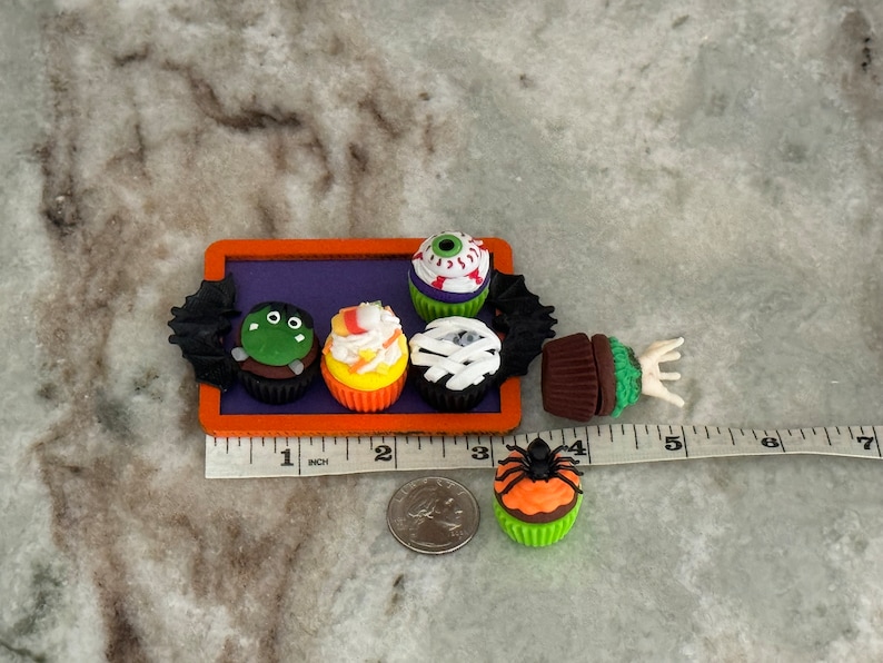 Op de afbeelding: Een set van zes Halloween-thema cupcakes met verschillende griezelige decoraties, waaronder een oog, een spin, een monster en snoep ma&iuml;s. De cupcakes zijn gerangschikt op een paarse dienblad met een zwart vleermuisontwerp. De dienblad staat op een wit aanrecht met een liniaal voor schaal.