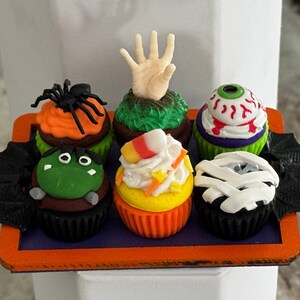 Op de afbeelding: Zes Halloween-cupcakes versierd met verschillende griezelige figuren en ontwerpen. De cupcakes zijn gerangschikt in een ronde oranje schaal met zwarte vleermuisvleugels aan beide kanten.